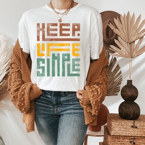 May include: White t-shirt with a graphic print that says "KEEP LIFE SIMPLE" in a layered, colorful design. The shirt is worn by a person with a brown cardigan and blue jeans.