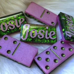 May include: Five dominoes with green glitter and a pink base. Four of the dominoes have the name "Josie" written on them in pink letters. One domino has two dots on one side and one dot on the other side.