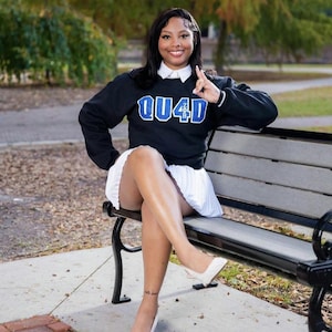 Puede incluir: Una sudadera negra con el texto "QU4D" en azul y blanco, combinada con una falda plisada blanca y tacones blancos. La persona está sentada en un banco del parque.