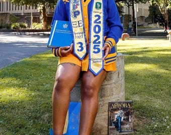 May include: A person wearing a blue and yellow cardigan, a white collared shirt, and a tie, with two graduation stoles featuring the text Sigma Gamma Rho, Spring 2023, and Class of 2025 Criminal Justice. They hold a blue book and wear blue heels.