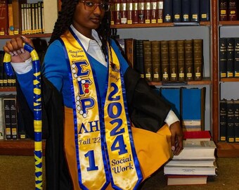 May include: A graduation ensemble featuring a black mortarboard cap, a blue long-sleeved shirt, and a gold skirt. Two gold satin stoles with blue trim display the text Alaysia Belmo, ΣΓΡ, ΛΗ, Fall '22, 1, Class of 2024, and Social Work. A blue and gold cane is held.