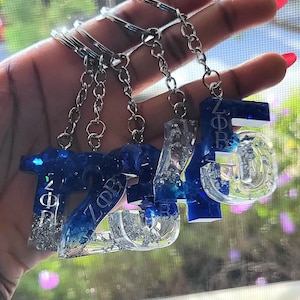 May include: Five blue and silver glitter resin keychains with silver key rings. Each keychain is a number, 1, 2, 3, 4, and 5. The number 5 has the letters "ZΦB" on it.