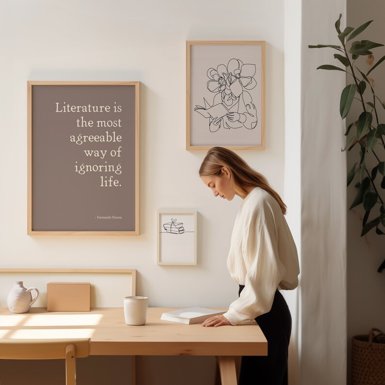 Puede incluir: Una mujer se encuentra de pie frente a una pared con tres impresiones enmarcadas. La impresi&oacute;n de la izquierda presenta una cita sobre literatura de Fernando Pessoa. La impresi&oacute;n del medio presenta un simple dibujo de l&iacute;nea de una pila de libros. La impresi&oacute;n de la derecha presenta un dibujo de l&iacute;nea de una mujer con flores que le crecen de la cabeza. La mujer de la imagen est&aacute; sosteniendo un libro y lo est&aacute; mirando.