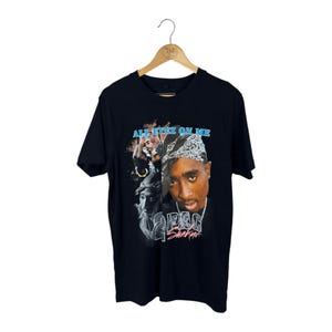 May include: Black t-shirt featuring a graphic of Tupac Shakur with the text "All Eyez On Me" in blue and "2Pac Shakur" in pink. The design includes images of Tupac and other figures. The shirt is hanging on a wooden hanger.