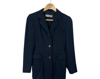 Puede incluir: Un blazer negro con solapa de muesca, cierre de tres botones y dos bolsillos delanteros. El blazer está colgado de una percha de madera. La etiqueta dice "JIL SANDER".