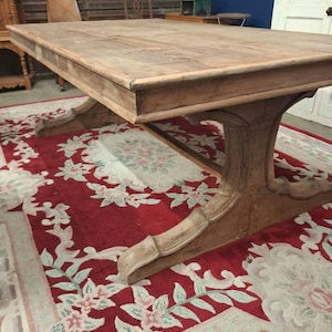 May include: A large rectangular wooden dining table with a natural light brown finish. The table features a thick, flat tabletop surface and is supported by two substantial, curved pedestal legs with decorative carved detailing at the base.