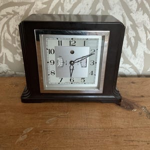 May include: A dark brown mantel clock with a square silver-toned face, black Arabic numerals, and two thin black hands. The clock features a metallic silver frame surrounding the dial, with small secondary indicators visible on the left and right.