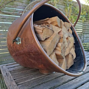 May include: A copper log holder with a black interior and a wooden handle. The holder is filled with stacked firewood.