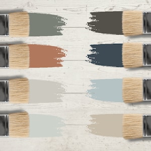 May include: Eight paintbrushes with wooden handles and natural bristles are arranged on a white wooden surface. Each brush displays a swatch of paint in a different color, including shades of green, brown, blue, and beige. The image showcases a color palette.