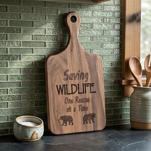 May include: A wooden chopping board with a handle, engraved with the words "Saving WILDLIFE One Rescue at a Time" and two bear illustrations. The board is brown and sits on a dark countertop, with a small ceramic bowl and utensils in the background.