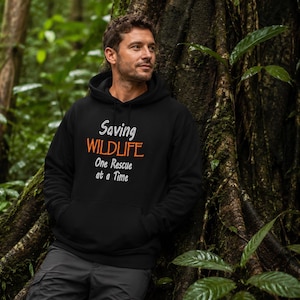 May include: A black hooded sweatshirt featuring the white text "Saving" above the orange text "WILDLIFE" and the white text "One Rescue at a Time" printed on the chest. The garment includes a front kangaroo pocket and a drawstring hood.