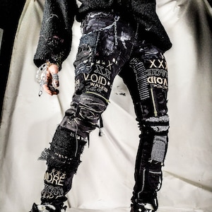 Model showing a black skinny pair with various abstract embroideries and words such as void walker and more riot with abstract symbols, black, white and grey patchwork, zippers, studs, splashes of gray and black paint and a chain attached to pocket.