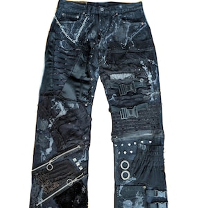 Könnte beinhalten: Schwarze Jeans im Patchwork-Design. Die Hose hat verschiedene Texturen, darunter Reißverschlüsse, Schnallen und eine Kette. Weißes Farbspritzer setzen Kontraste. Der Stil ist edgy und einzigartig.