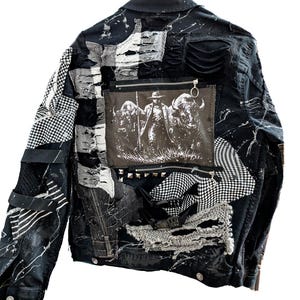 Peut inclure: Une veste en jean noire au style usé avec des éclaboussures de peinture blanche, des empiècements en tissu effiloché et un panneau central rectangulaire représentant une personne chapeautée entre deux buffles. Inclut des clous et fermetures.