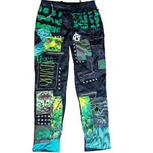 May include: Black denim pants with multiple patches and embellishments. The pants feature green and black graphics, including a face, mushrooms, and skulls. Silver studs and zippers add detail. The pants have a unique, distressed design.
