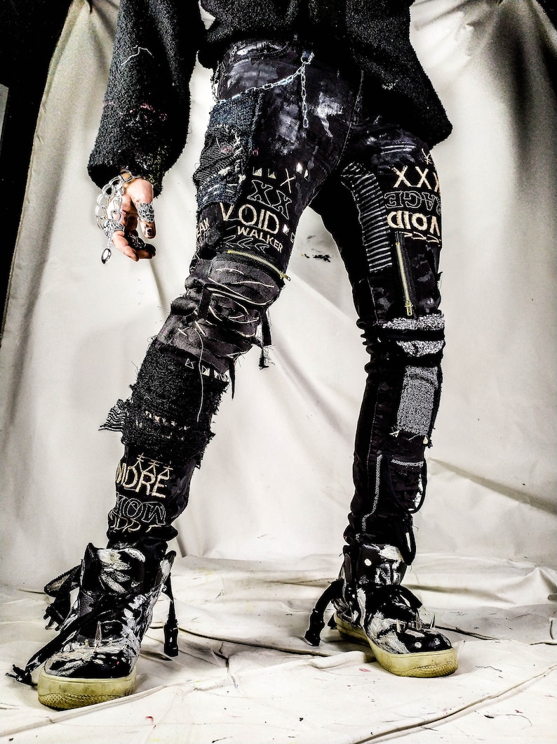 Model showing a black skinny pair with various abstract embroideries and words such as void walker and more riot with abstract symbols, black, white and grey patchwork, zippers, studs, splashes of gray and black paint and a chain attached to pocket.