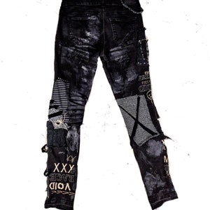 Flat laid black skinny pair with various abstract embroideries and words such as void walker and more riot with abstract symbols, black, white and grey patchwork, zippers, studs, splashes of gray and black paint and a chain attached to pocket.