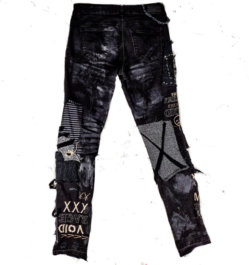Flat laid black skinny pair with various abstract embroideries and words such as void walker and more riot with abstract symbols, black, white and grey patchwork, zippers, studs, splashes of gray and black paint and a chain attached to pocket.