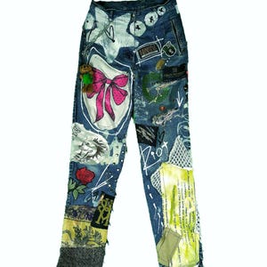 May include: Blue denim jeans with various patches, including a pink bow, a rose, and text. The jeans have a distressed look with paint splatters and a teddy bear charm. The jeans are a unique, artistic fashion statement.