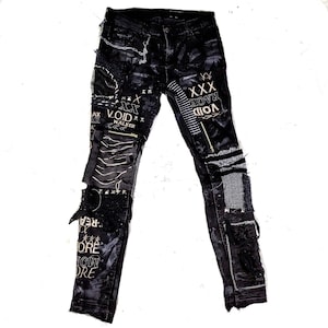 Flat laid black skinny pair with various abstract embroideries and words such as void walker and more riot with abstract symbols, black, white and grey patchwork, zippers, studs, splashes of gray and black paint and a chain attached to pocket.