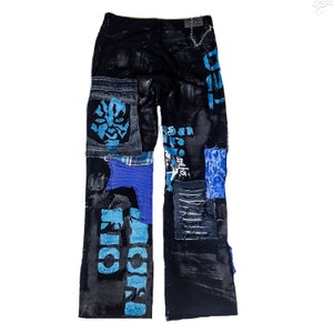 Flat laid black pants with anime prints, colorful patchwork in multiple blue shades, ripped patches, zipper, eyelets, studs and abstract painting with a blue snake on the front.