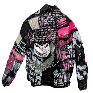 May include: A black jacket featuring a patchwork design with pink and white accents. The garment includes distressed fabric, a white patch with a jagged grin, a checkered pattern, skull motifs, and a silver chain detail across the front surface.