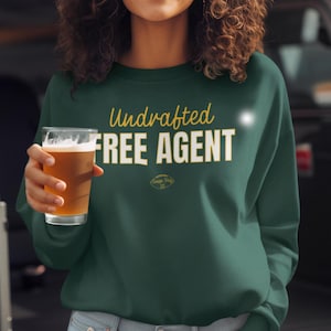 May include: Dark green sweatshirt with the words "Undrafted FREE AGENT" in gold and white lettering. The sweatshirt also features a small graphic and is paired with light blue jeans. A glass of beer is held in the hand.