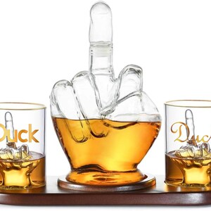 May include: A clear glass decanter and two matching glasses, all shaped like a hand giving the middle finger. The decanter is filled with amber liquid and sits on a wooden tray. The word "Duck" is written on the glasses.