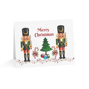 May include: A white greeting card featuring the text Merry Christmas in red script. The design includes two nutcracker figures in green and red uniforms, a central green Christmas tree, two candy canes, and colorful ornaments at the base.