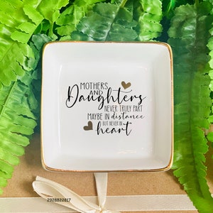Puede incluir: Plato de cerámica blanca para joyas con borde dorado. El plato tiene una inscripción en negro que dice "Madres e hijas nunca se separan realmente Tal vez en la distancia Pero nunca en el corazón" con dos corazones dorados.