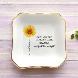 Puede incluir: Plato de cerámica blanco con borde dorado, con una ilustración de girasol y el texto "EVEN ON THE DARKEST DAYS, stand tall and find the sunlight." El plato es cuadrado con bordes redondeados.