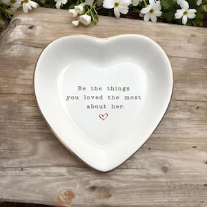 May include: A white heart-shaped trinket dish with a gold rim. The dish has the quote "Be the things you loved the most about her." in black text, with a small red heart below. The dish is on a wooden surface.