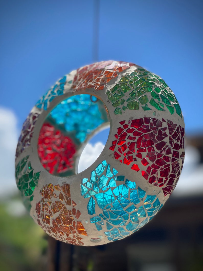 May include: A colorful mosaic glass sphere hanging from a string. The sphere is made of small pieces of glass in shades of red, orange, yellow, green, blue, and white. The sphere has a hole in the center, making it suitable for hanging.