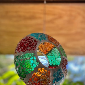 May include: A colorful mosaic glass sphere with a hole in the center, hanging from a ceiling. The sphere is made of small, irregular pieces of glass in shades of red, green, blue, and orange.