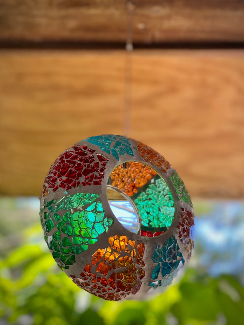 May include: A colorful mosaic glass sphere with a hole in the center, hanging from a ceiling. The sphere is made of small, irregular pieces of glass in shades of red, green, blue, and orange.