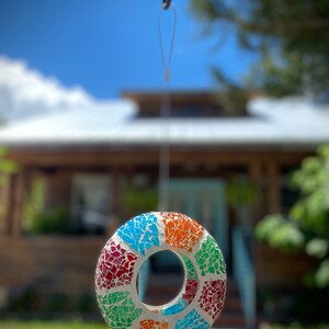 May include: A colorful mosaic bird feeder with a round shape and a hole in the center, hanging from a hook against a blurred background of a house and green grass.