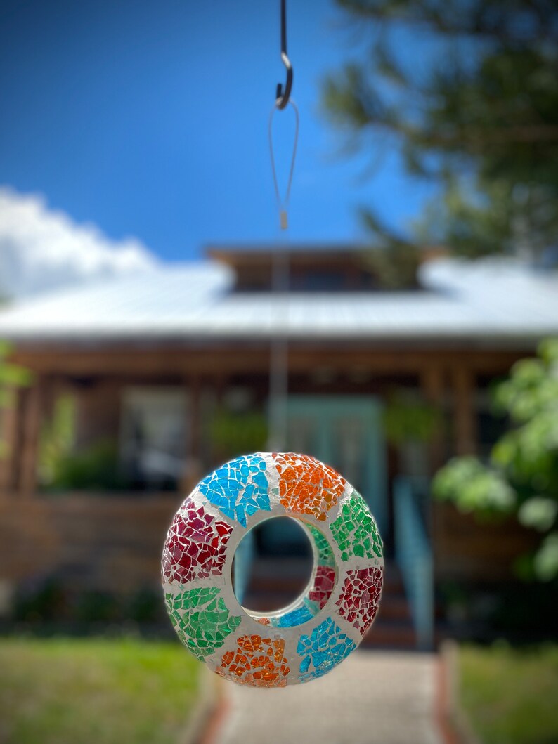 May include: A colorful mosaic bird feeder with a round shape and a hole in the center, hanging from a hook against a blurred background of a house and green grass.