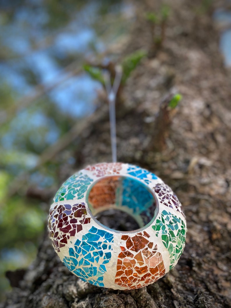 May include: A round, mosaic-tiled, hanging candle holder with a teal, brown, and white color scheme. The holder is made of small, irregular pieces of glass.