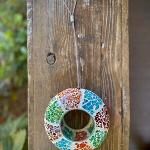 May include: A colorful mosaic bird feeder with a round shape and a hole in the center. It is hanging from a white cord.