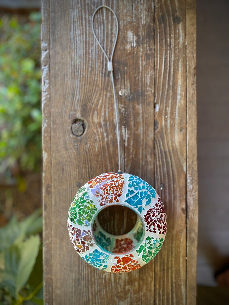 May include: A colorful mosaic bird feeder with a round shape and a hole in the center. It is hanging from a white cord.