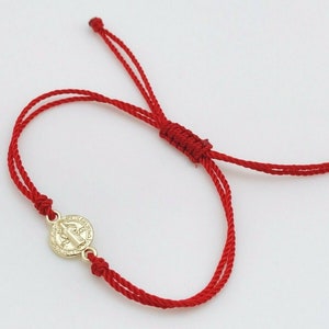 May include: Red string bracelet featuring a gold-colored medallion. The medallion has a religious design. The bracelet is adjustable with sliding knots. The cord is a vibrant red color.