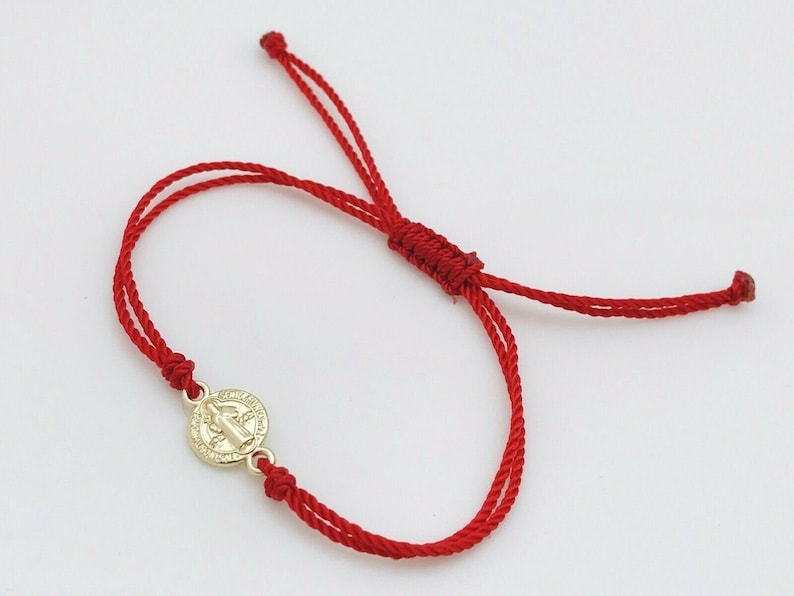 May include: Red string bracelet featuring a gold-colored medallion. The medallion has a religious design. The bracelet is adjustable with sliding knots. The cord is a vibrant red color.