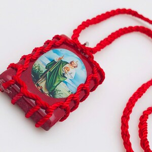 May include: A red pendant necklace featuring an image of a religious figure. The pendant is rectangular with a red border and a braided red cord. The image is of a man in green and white robes. The necklace is on a white background.