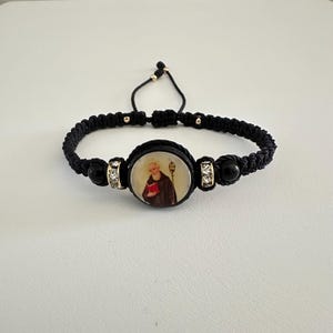 Peut inclure: Bracelet en macramé noir orné d'un pendentif ovale représentant une figure religieuse. Le bracelet est agrémenté de perles noires et de détails dorés. La conception réglable assure un ajustement confortable.
