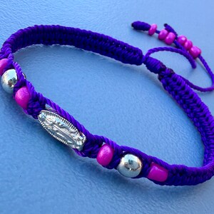 Peut inclure: Bracelet tressé violet avec des perles roses et un charme argenté. Le charme représente une image religieuse.