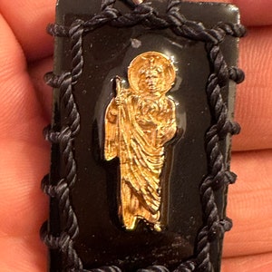 May include: A black pendant with a gold image of a saint inside. The pendant is framed with black rope and has a silver loop at the top.