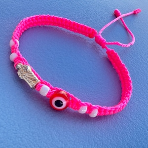 Peut inclure: Bracelet tressé rose avec un charme argenté, des perles blanches et un œil de protection rouge.