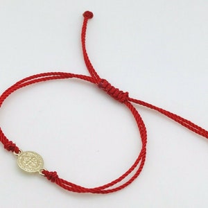 May include: A red cord bracelet with a gold-colored circular charm. The charm features an embossed design. The bracelet is adjustable with sliding knots and is displayed on a white background.
