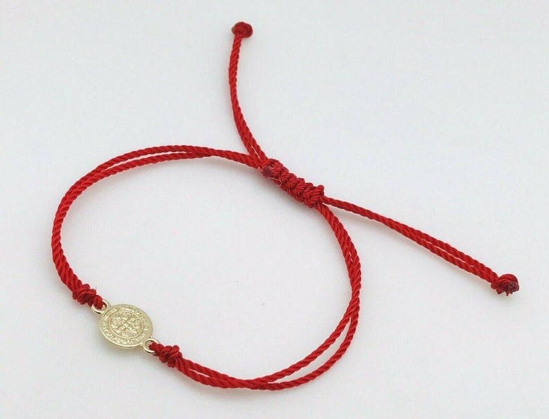 May include: A red cord bracelet with a gold-colored circular charm. The charm features an embossed design. The bracelet is adjustable with sliding knots and is displayed on a white background.