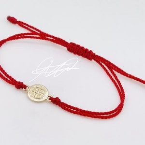 May include: Red string bracelet with a gold-colored medallion. The medallion features a detailed design. The bracelet is adjustable with a sliding knot closure. The design is simple and elegant.